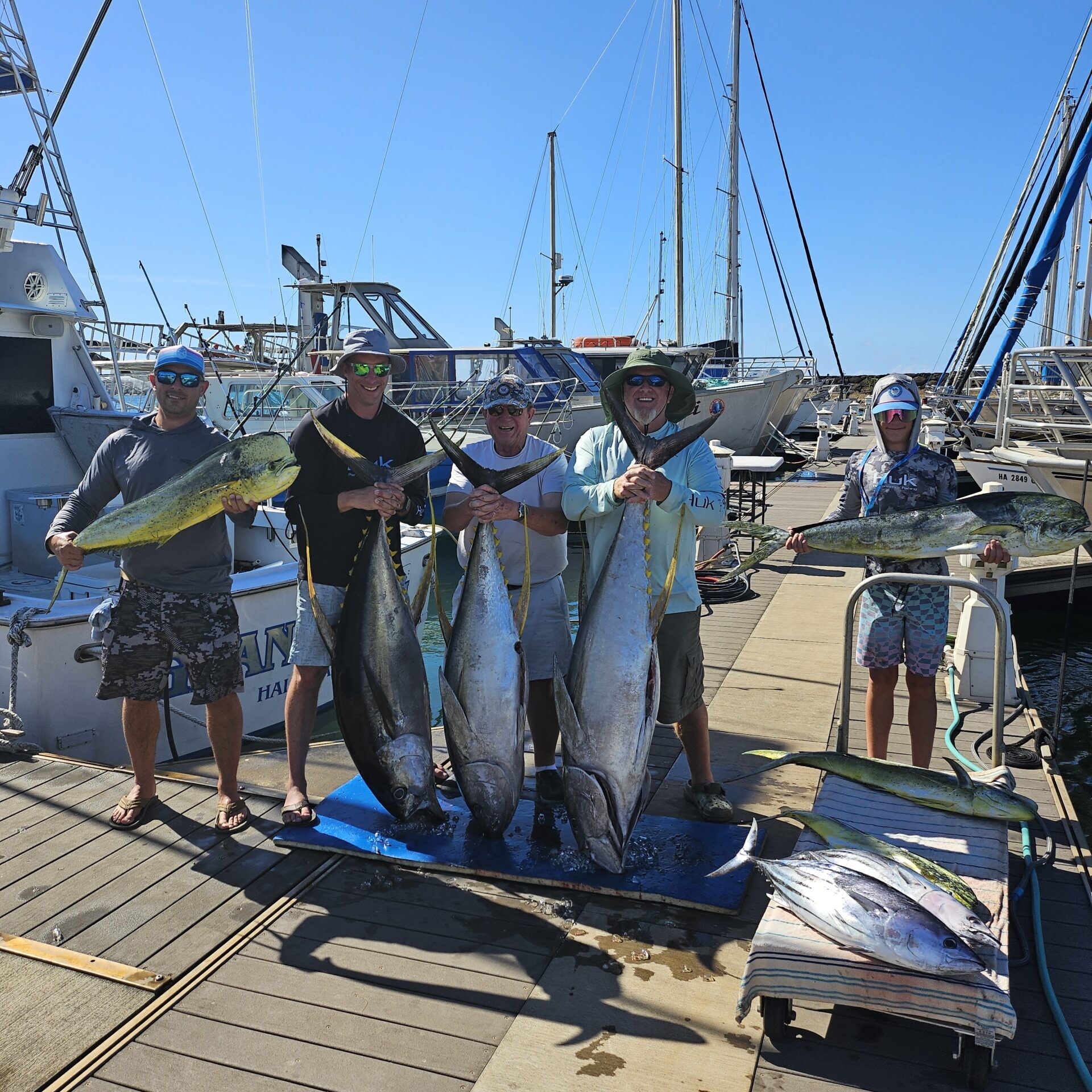 Four men proudly hold large fish at a marina dock with boats in the background.
